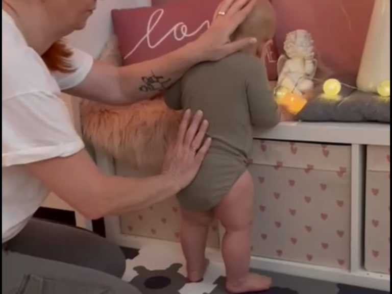 Eine Erwachsene hilft einem Baby, das am Möbel steht, mit einer sanften Berührung.